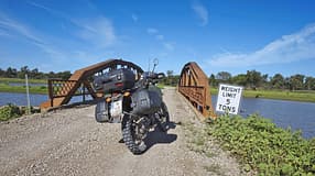 Das Foto zeigt ein Reiseenduro‑Motorrad mit montierten Seitenkoffern und Topcase auf einer Schotterpiste vor einer kleinen stählernen Brücke über ein Gewässer. Stollenreifen, hohe Bodenfreiheit und Gepäcksystem betonen den Offroad‑Einsatz. Am Brückeneingang steht ein Schild mit der Aufschrift „WEIGHT LIMIT 5 TONS“ in ländlicher Umgebung unter blauem Himmel.