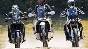 Die Aufnahme zeigt drei Reiseenduros in der Frontansicht: CFMOTO 1000MT-X, Ducati DesertX und Honda CRF1100L Africa Twin. Die Adventure-Bikes fahren nebeneinander auf einer Schotterstraße, mit hohen Windschilden und Handprotektoren gut erkennbar. Im leichten Offroad-Einsatz wirbeln sie Staub auf, die Szene wirkt wie ein Vergleichslauf im Gelände.