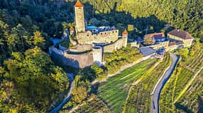 Die Luftaufnahme zeigt eine mittelalterliche Burganlage auf einem bewaldeten Hügel mit markantem Bergfried und mehreren Türmen mit roten Zeltdächern. Massive Wehrmauern umschließen Innenhöfe und Gebäude; am Hang liegen terrassierte Weinberge, daneben verläuft eine kurvige Zufahrtsstraße.