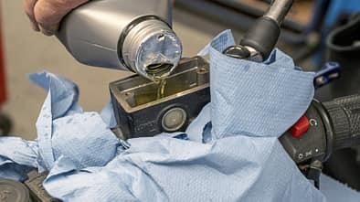 Bremsflüssigkeitswechsel an einem Motorrad in einer Werkstatt. Eine Hand füllt Bremsflüssigkeit in den Vorratsbehälter am Lenker, der mit blauen Tüchern abgedeckt ist.