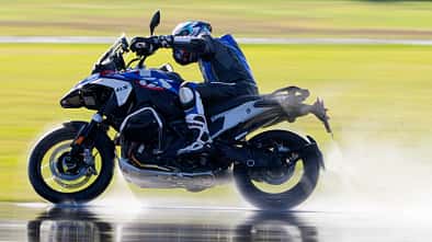 Reiseenduro-Motorrad mit Fahrer auf nasser Fahrbahn, dynamische Fahrszene mit Wasserfontäne.