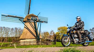Motorradfahrer auf Landstraße vor Windmühle und Windkraftanlage, ländliche Umgebung, blauer Himmel.