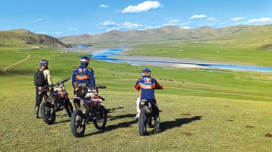 Enduro, KTM, Motorrad, Offroad, Abenteuer, Landschaft, Fluss, Mongolei, Motorradtour, Fernreise Drei Enduro-Motorradfahrer mit KTM-Bekleidung stehen mit ihren Motorrädern auf einer weiten Graslandschaft mit Fluss und Bergen im Hintergrund.