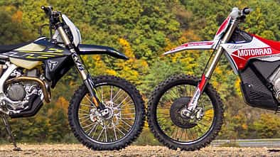 Enduro, Motorrad, Geländemotorrad, Offroad, Motorradtechnik, Windenergie, Wald, Motorsport, Zweirad, Natur, Triumph TF 450-E, Stark Varg EX Zwei Enduro-Motorräder, die Stark Varg EX und die Triumph TF 450-E parken frontal gegenüber im Gelände vor bewaldetem Hintergrund und Windrädern, sodass sich die Vorderreifen berühren. Aufnahme aus der Seitenansicht.