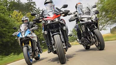 Suzuki GSX-S1000 GX, Honda CB 1000 GT, Kawasaki Versys 1100 SE, Tourer, Motorradvergleich, Straßenmotorrad, Landstraße, Fahrtest, 2026, Reiseenduro Drei Motorräder – Suzuki GSX-S1000 GX, Honda CB 1000 GT und Kawasaki Versys 1100 SE – fahren nebeneinander auf einer Landstraße. Die Aufnahme zeigt die Motorräder frontal in Bewegung mit Fahrern in voller Schutzkleidung. Die Umgebung ist grün und leicht verschwommen, was die Dynamik der Fahrt unterstreicht.