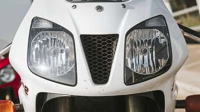 Die Detailaufnahme zeigt die Frontverkleidung eines Honda‑Sportmotorrads mit deutlich sichtbarem Honda‑Schriftzug. Zu sehen sind zwei große Doppelscheinwerfer und ein zentraler, dreieckiger Lufteinlass mit Wabengitter; unten sind die Standrohre der Telegabel sowie die Blinker erkennbar.