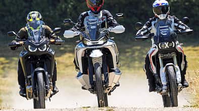 Die Aufnahme zeigt drei Reiseenduros in der Frontansicht: CFMOTO 1000MT-X, Ducati DesertX und Honda CRF1100L Africa Twin. Die Adventure-Bikes fahren nebeneinander auf einer Schotterstraße, mit hohen Windschilden und Handprotektoren gut erkennbar. Im leichten Offroad-Einsatz wirbeln sie Staub auf, die Szene wirkt wie ein Vergleichslauf im Gelände.