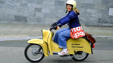 Das Foto zeigt einen gelben Kleinroller im klassischen Design, der von einer Fahrerin mit Helm auf einer städtischen Straße bewegt wird. Auf der Seitenverkleidung steht „KULTURGUT TRIFFT ZUKUNFT“, was auf eine Elektro‑Nachrüstung mit rückrüstbarem E‑Antrieb hindeutet. Die Szene ist seitlich aufgenommen vor einer Steinmauer und umfasst Fahrbahn, Bordstein und Gehweg.