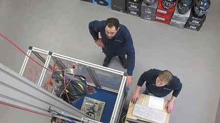 Zwei Personen in einem Labor oder Prüfraum, die einen Motorradhelm an einem Prüfstand testen. Im Hintergrund sind mehrere Motorradhelme aufgestapelt.