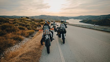 Adventure-Motorräder am Straßenrand mit Blick über eine Landschaft und Wasser, Fahrer mit Helmen.