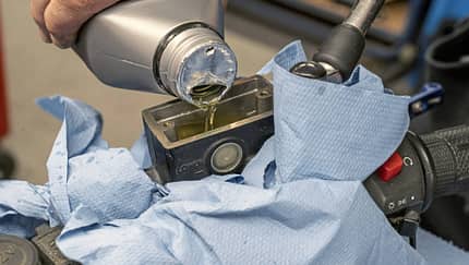 Bremsflüssigkeitswechsel an einem Motorrad in einer Werkstatt. Eine Hand füllt Bremsflüssigkeit in den Vorratsbehälter am Lenker, der mit blauen Tüchern abgedeckt ist.