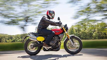 Fantic Caballero 700 Motorrad in Fahrt auf Landstraße, seitliche Ansicht, Fahrer mit Helm und Lederjacke, unscharfer Hintergrund durch Mitzieheffekt.