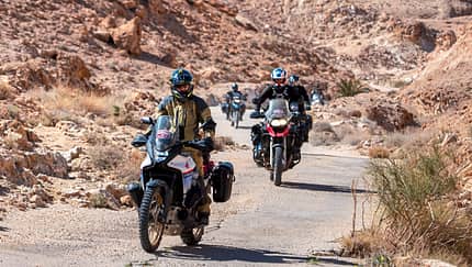 Zwei Motorradfahrer fahren auf einer schlecht geteerten Straße durch eine sandige Gegend