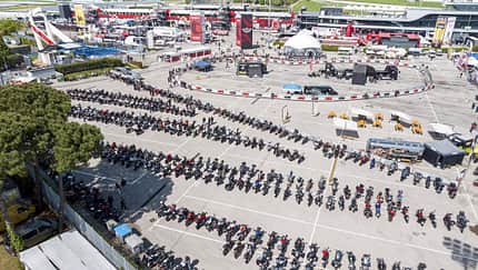 Luftaufnahme eines großen Motorrad-Events mit zahlreichen Motorrädern auf einem Parkplatz und Event-Areal mit Messeständen.