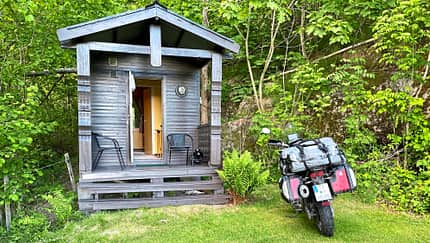 Reiseenduro Moto Guzzi V85 TT mit Gepäck vor einer kleinen Holzhütte im Grünen. Die Szene zeigt eine typische Motorradtour-Unterkunft.