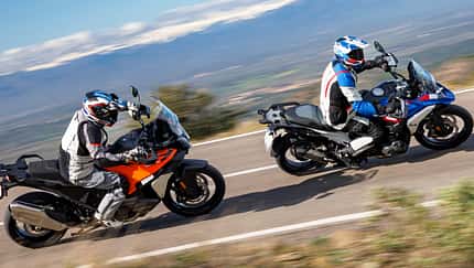 Zwei Reiseenduro-Motorräder, BMW R 1300 GS Trophy und KTM 1390 Super Adventure S Evo, fahren nebeneinander in Kurvenfahrt auf einem Schotterweg. Beide Fahrer tragen Motorradbekleidung und Helme. Im Hintergrund sind Bäume, Steine und Berge zu sehen.