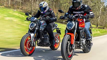 Zwei Ducati Monster Motorräder, neu und alt, fahren nebeneinander auf einer Landstraße. Beide Fahrer tragen Motorradbekleidung und Helme. Die Aufnahme zeigt einen direkten Vergleich der beiden Modellgenerationen in Bewegung.