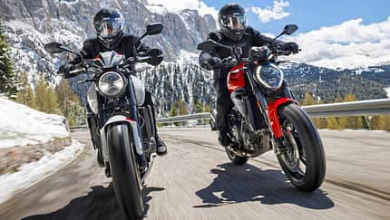 Ducati Monster+, Triumph Trident 800, Alpen Masters 2026, Mid-Naked Bikes, Alpen, Motorradtest, Naked Bike Zwei Mid-Naked-Motorräder beim Alpenmaster 2024 auf einer Passstraße in den Alpen, beide Fahrer mit Motorradbekleidung und Helmen, verschneite Berge im Hintergrund.