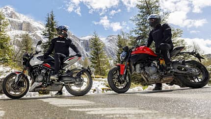 Ducati Monster+, Triumph Trident 800, Alpen Masters 2026, Mid-Naked Bikes, Alpen, Motorradtest, Naked Bike Zwei Mid-Naked-Motorräder mit Fahrern auf Alpenstraße, Schneereste am Straßenrand, Gebirgslandschaft im Hintergrund.