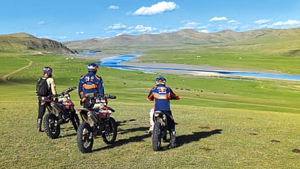 Enduro, KTM, Motorrad, Offroad, Abenteuer, Landschaft, Fluss, Mongolei, Motorradtour, Fernreise Drei Enduro-Motorradfahrer mit KTM-Bekleidung stehen mit ihren Motorrädern auf einer weiten Graslandschaft mit Fluss und Bergen im Hintergrund.