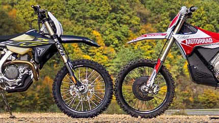 Zwei Enduro-Motorräder, die Stark Varg EX und die Triumph TF 450-E parken frontal gegenüber im Gelände vor bewaldetem Hintergrund und Windrädern, sodass sich die Vorderreifen berühren. Aufnahme aus der Seitenansicht.
