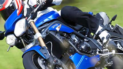 Triumph Speed Triple, Naked Bike, Motorrad, Straßenmotorrad, Dreizylinder, Schräglage, Landstraße, Fahrdynamik, Actionaufnahme Eine Triumph Speed Triple in blauer Lackierung wird in Schräglage auf einer Landstraße gefahren. Der Fahrer trägt schwarze Motorradbekleidung und einen rot-weißen Helm.