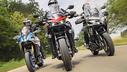Drei Motorräder – Suzuki GSX-S1000 GX, Honda CB 1000 GT und Kawasaki Versys 1100 SE – fahren nebeneinander auf einer Landstraße. Die Aufnahme zeigt die Motorräder frontal in Bewegung mit Fahrern in voller Schutzkleidung. Die Umgebung ist grün und leicht verschwommen, was die Dynamik der Fahrt unterstreicht.