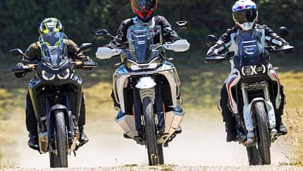 CFMOTO 1000MT-X, Ducati DesertX, Honda CRF1100L Africa Twin, Reiseenduro, Adventure-Bike, Vergleichstest, Offroad-Fahrt, Schotterstraße, Dreiervergleich, Motorrad Die Aufnahme zeigt drei Reiseenduros in der Frontansicht: CFMOTO 1000MT-X, Ducati DesertX und Honda CRF1100L Africa Twin. Die Adventure-Bikes fahren nebeneinander auf einer Schotterstraße, mit hohen Windschilden und Handprotektoren gut erkennbar. Im leichten Offroad-Einsatz wirbeln sie Staub auf, die Szene wirkt wie ein Vergleichslauf im Gelände.