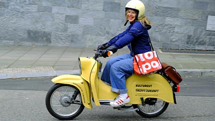 Das Foto zeigt einen gelben Kleinroller im klassischen Design, der von einer Fahrerin mit Helm auf einer städtischen Straße bewegt wird. Auf der Seitenverkleidung steht „KULTURGUT TRIFFT ZUKUNFT“, was auf eine Elektro‑Nachrüstung mit rückrüstbarem E‑Antrieb hindeutet. Die Szene ist seitlich aufgenommen vor einer Steinmauer und umfasst Fahrbahn, Bordstein und Gehweg.