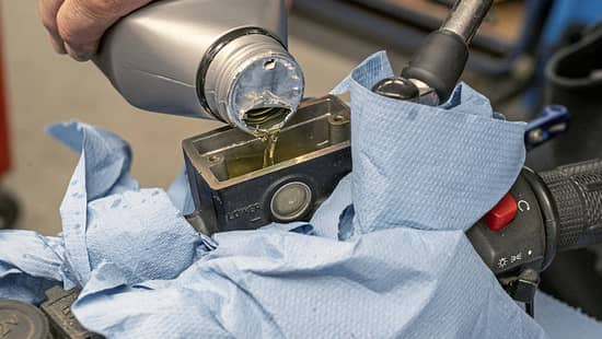 Bremsflüssigkeitswechsel an einem Motorrad in einer Werkstatt. Eine Hand füllt Bremsflüssigkeit in den Vorratsbehälter am Lenker, der mit blauen Tüchern abgedeckt ist.