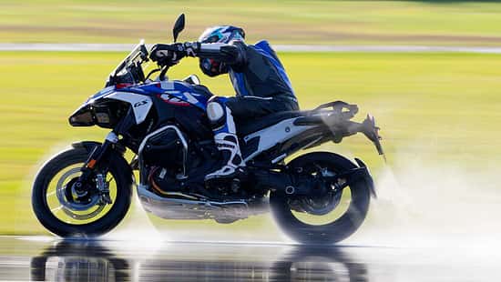 Reiseenduro-Motorrad mit Fahrer auf nasser Fahrbahn, dynamische Fahrszene mit Wasserfontäne.