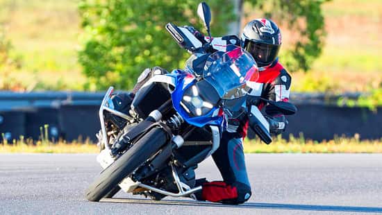 Motorradfahrer in voller Schräglage auf einer asphaltierten Rennstrecke, Motorrad mit großem Windschild und Sturzbügeln, sportliche Schutzkleidung, sommerliche Umgebung.