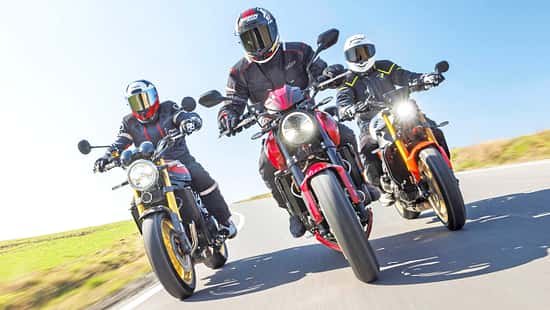 Drei Naked Bikes (Kawasaki Z900RS SE, Triumph Trident 800, Yamaha XSR900) fahren nebeneinander auf einer Landstraße, aufgenommen aus der Froschperspektive. Die Fahrer tragen Motorradbekleidung und Integralhelme, im Hintergrund ist ein Windrad zu sehen.