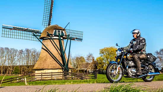 Motorradfahrer auf Landstraße vor Windmühle und Windkraftanlage, ländliche Umgebung, blauer Himmel.