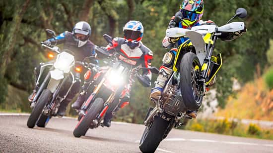 Drei Supermoto-Motorräder, Ducati Hypermotard 698 Mono RVE, Husqvarna 701 Supermoto und Suzuki DR-Z4SM, fahren auf einer Landstraße. Der Fahrer auf der Husqvarna 701 Supermoto macht einen Wheelie, die anderen beiden fahren nebeneinander. Alle Fahrer tragen Motorradschutzkleidung und Helme. Die Szene spielt sich auf einer kurvigen Straße im Grünen ab.