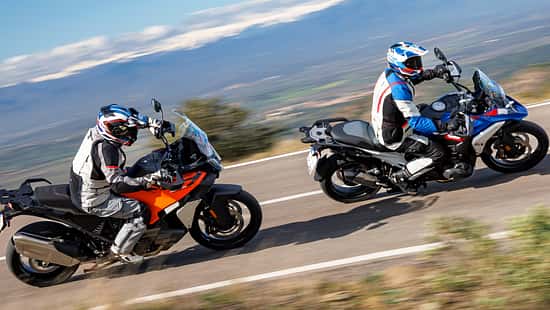 Zwei Reiseenduro-Motorräder, BMW R 1300 GS Trophy und KTM 1390 Super Adventure S Evo, fahren nebeneinander in Kurvenfahrt auf einem Schotterweg. Beide Fahrer tragen Motorradbekleidung und Helme. Im Hintergrund sind Bäume, Steine und Berge zu sehen.