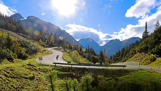 Motorradfahrer in Kurve auf Passstraße am Hahntennjoch in alpiner Landschaft bei Sonnenschein.
