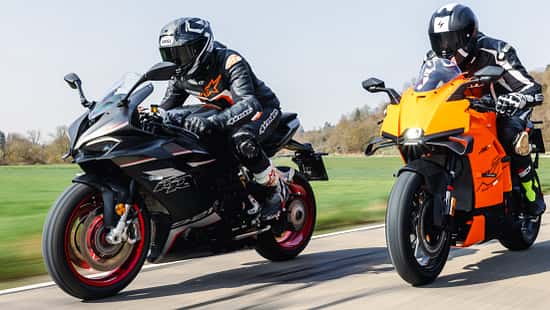 Zwei Sportmotorräder, QJ Motor SRK 921 RR und KTM 990 RC R, werden auf einer Landstraße gefahren. Beide Fahrer tragen Motorradbekleidung und Integralhelme. Die Aufnahme zeigt die Motorräder in voller Fahrt bei Tageslicht.