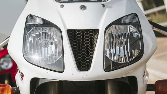 Die Detailaufnahme zeigt die Frontverkleidung eines Honda‑Sportmotorrads mit deutlich sichtbarem Honda‑Schriftzug. Zu sehen sind zwei große Doppelscheinwerfer und ein zentraler, dreieckiger Lufteinlass mit Wabengitter; unten sind die Standrohre der Telegabel sowie die Blinker erkennbar.