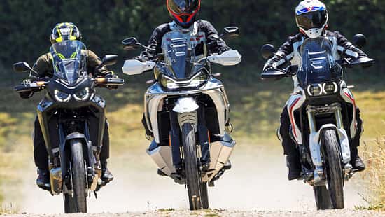 Die Aufnahme zeigt drei Reiseenduros in der Frontansicht: CFMOTO 1000MT-X, Ducati DesertX und Honda CRF1100L Africa Twin. Die Adventure-Bikes fahren nebeneinander auf einer Schotterstraße, mit hohen Windschilden und Handprotektoren gut erkennbar. Im leichten Offroad-Einsatz wirbeln sie Staub auf, die Szene wirkt wie ein Vergleichslauf im Gelände.