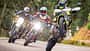 Drei Supermoto-Motorräder, Ducati Hypermotard 698 Mono RVE, Husqvarna 701 Supermoto und Suzuki DR-Z4SM, fahren auf einer Landstraße. Der Fahrer auf der Husqvarna 701 Supermoto macht einen Wheelie, die anderen beiden fahren nebeneinander. Alle Fahrer tragen Motorradschutzkleidung und Helme. Die Szene spielt sich auf einer kurvigen Straße im Grünen ab.