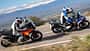 Zwei Reiseenduro-Motorräder, BMW R 1300 GS Trophy und KTM 1390 Super Adventure S Evo, fahren nebeneinander in Kurvenfahrt auf einem Schotterweg. Beide Fahrer tragen Motorradbekleidung und Helme. Im Hintergrund sind Bäume, Steine und Berge zu sehen.