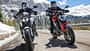 Ducati Monster+, Triumph Trident 800, Alpen Masters 2026, Mid-Naked Bikes, Alpen, Motorradtest, Naked Bike Zwei Mid-Naked-Motorräder beim Alpenmaster 2024 auf einer Passstraße in den Alpen, beide Fahrer mit Motorradbekleidung und Helmen, verschneite Berge im Hintergrund.