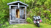 Reiseenduro Moto Guzzi V85 TT mit Gepäck vor einer kleinen Holzhütte im Grünen. Die Szene zeigt eine typische Motorradtour-Unterkunft.