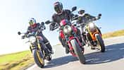 Drei Naked Bikes (Kawasaki Z900RS SE, Triumph Trident 800, Yamaha XSR900) fahren nebeneinander auf einer Landstraße, aufgenommen aus der Froschperspektive. Die Fahrer tragen Motorradbekleidung und Integralhelme, im Hintergrund ist ein Windrad zu sehen.