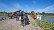 Das Foto zeigt ein Reiseenduro‑Motorrad mit montierten Seitenkoffern und Topcase auf einer Schotterpiste vor einer kleinen stählernen Brücke über ein Gewässer. Stollenreifen, hohe Bodenfreiheit und Gepäcksystem betonen den Offroad‑Einsatz. Am Brückeneingang steht ein Schild mit der Aufschrift „WEIGHT LIMIT 5 TONS“ in ländlicher Umgebung unter blauem Himmel.
