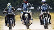 Die Aufnahme zeigt drei Reiseenduros in der Frontansicht: CFMOTO 1000MT-X, Ducati DesertX und Honda CRF1100L Africa Twin. Die Adventure-Bikes fahren nebeneinander auf einer Schotterstraße, mit hohen Windschilden und Handprotektoren gut erkennbar. Im leichten Offroad-Einsatz wirbeln sie Staub auf, die Szene wirkt wie ein Vergleichslauf im Gelände.